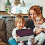 two babies and woman sitting on sofa while holding baby and watching on tablet